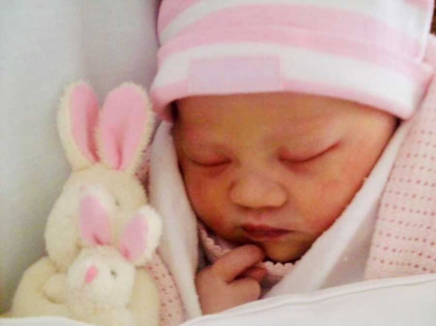 Baby girl with down syndrome lays asleep wrapped in pink blanket and hat beside small stuffed bunnies