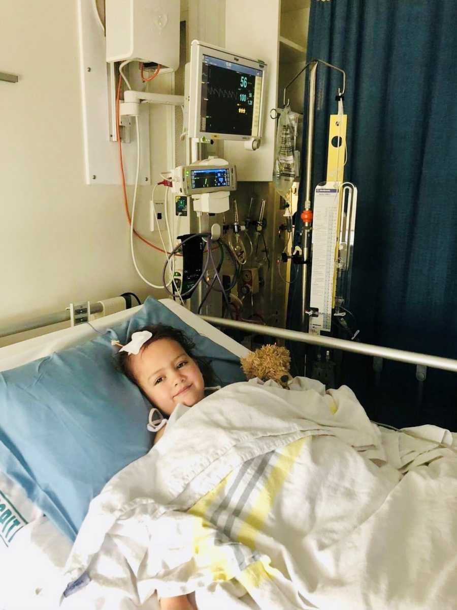 Little girl with mass on her brain lays in hospital bed smiling with stuffed animal at her side