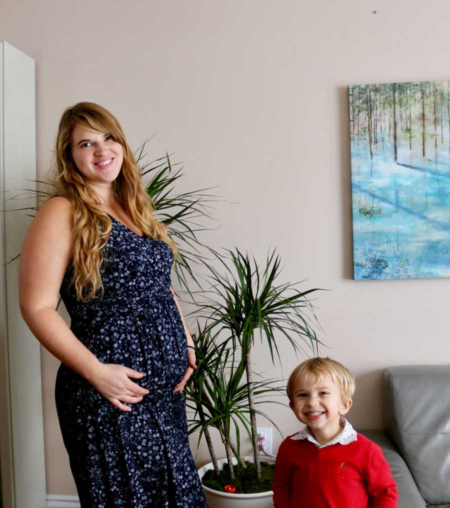 Pregnant woman stands in home holding her stomach as her son stands smiling next to her