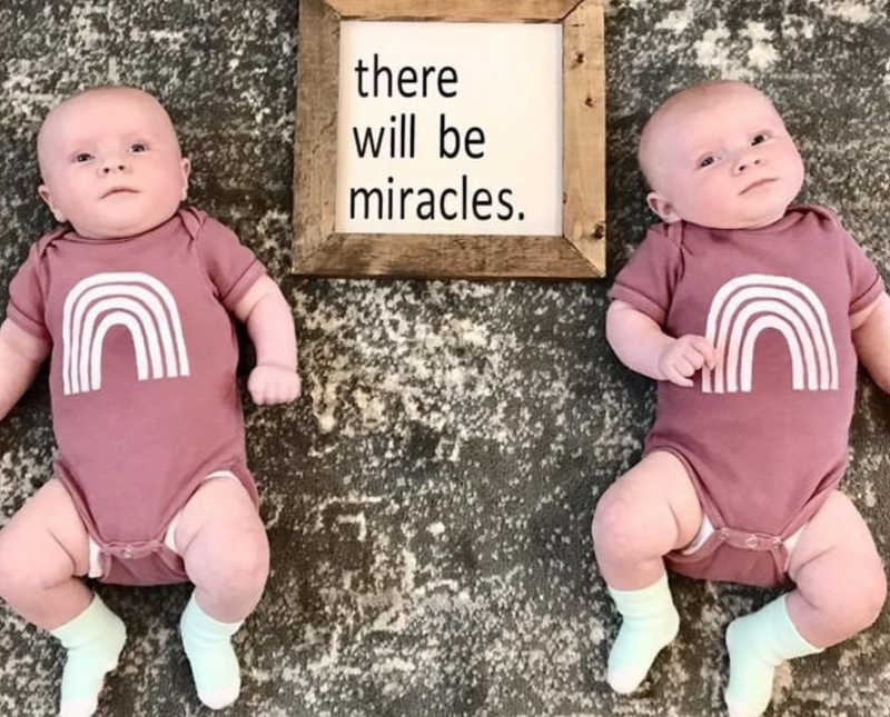 Twin babies lay in matching onesies with rainbows on it beside sign that says, "there will be miracles"