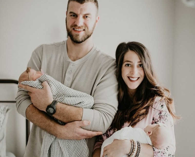Husband and wife stands smiling as they hold their newborn twins