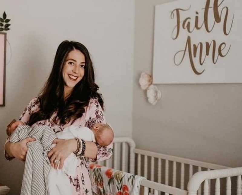 Mother stands holding newborn twins beside crib