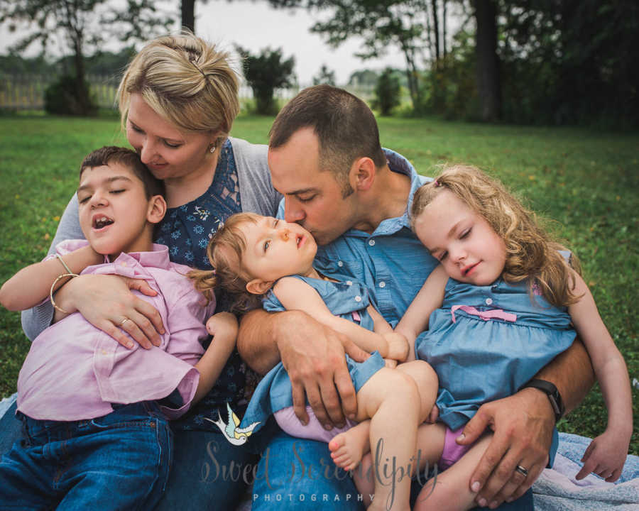 Husband and wife sit on ground outside with their three adopted children with special needs in their laps