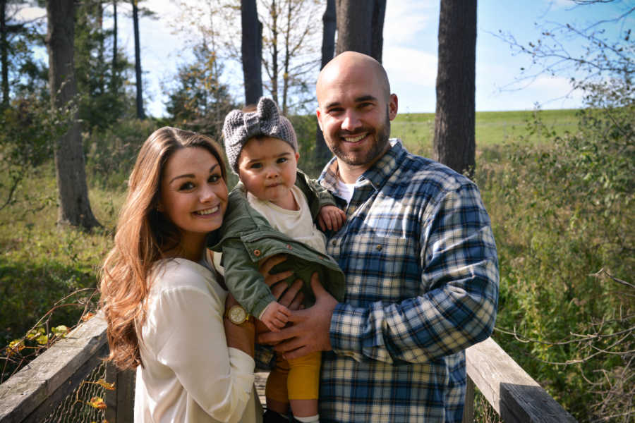 Husband and wife smile as they stand on wooden bridge outside holding their daughter