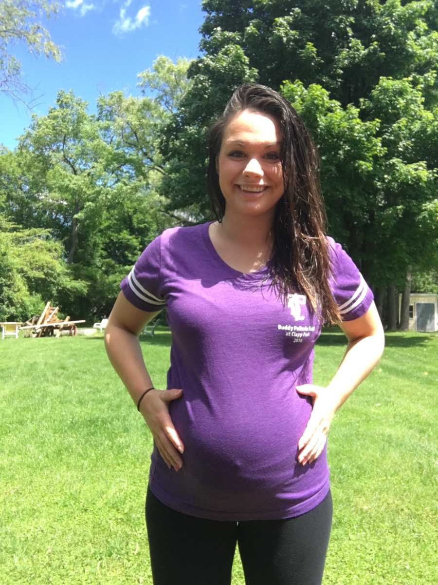 Pregnant woman stands outside smiling with hands on her stomach
