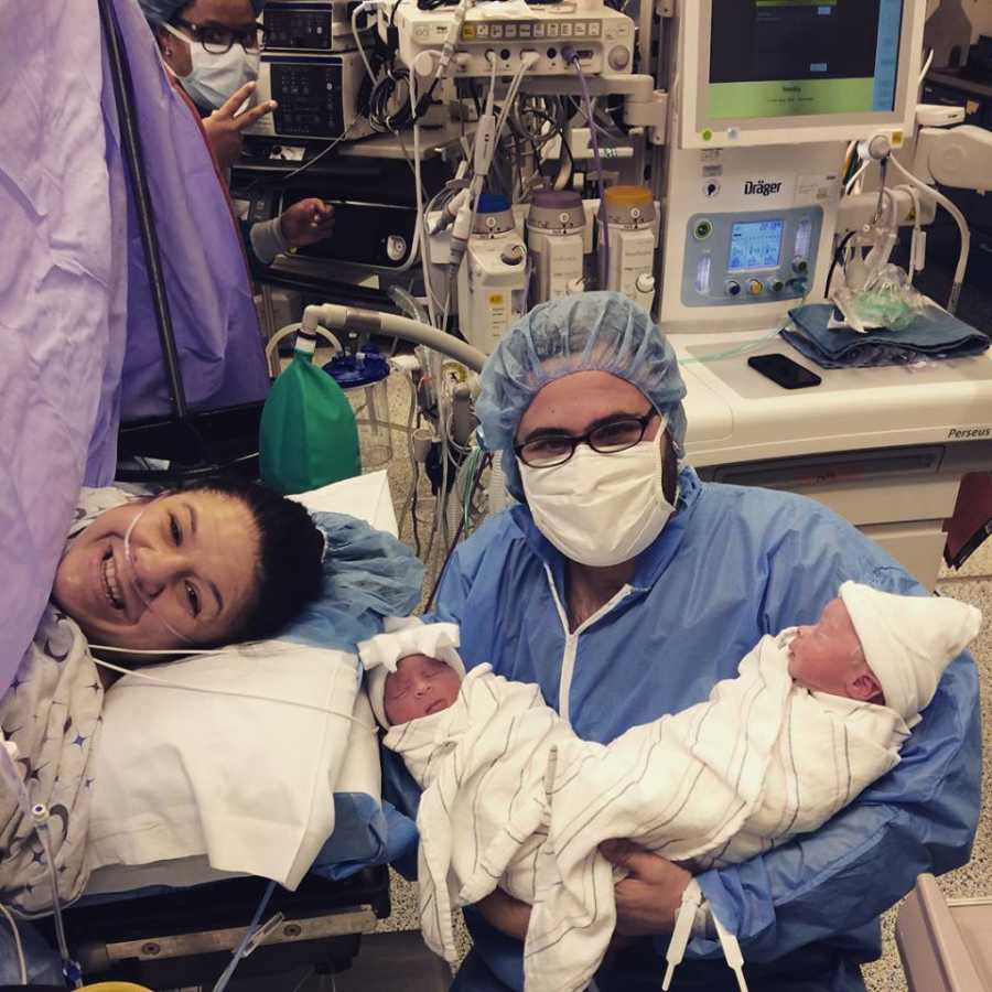 Mother who just had c-section lays on operating table smiling while her husband kneels beside her holding twins