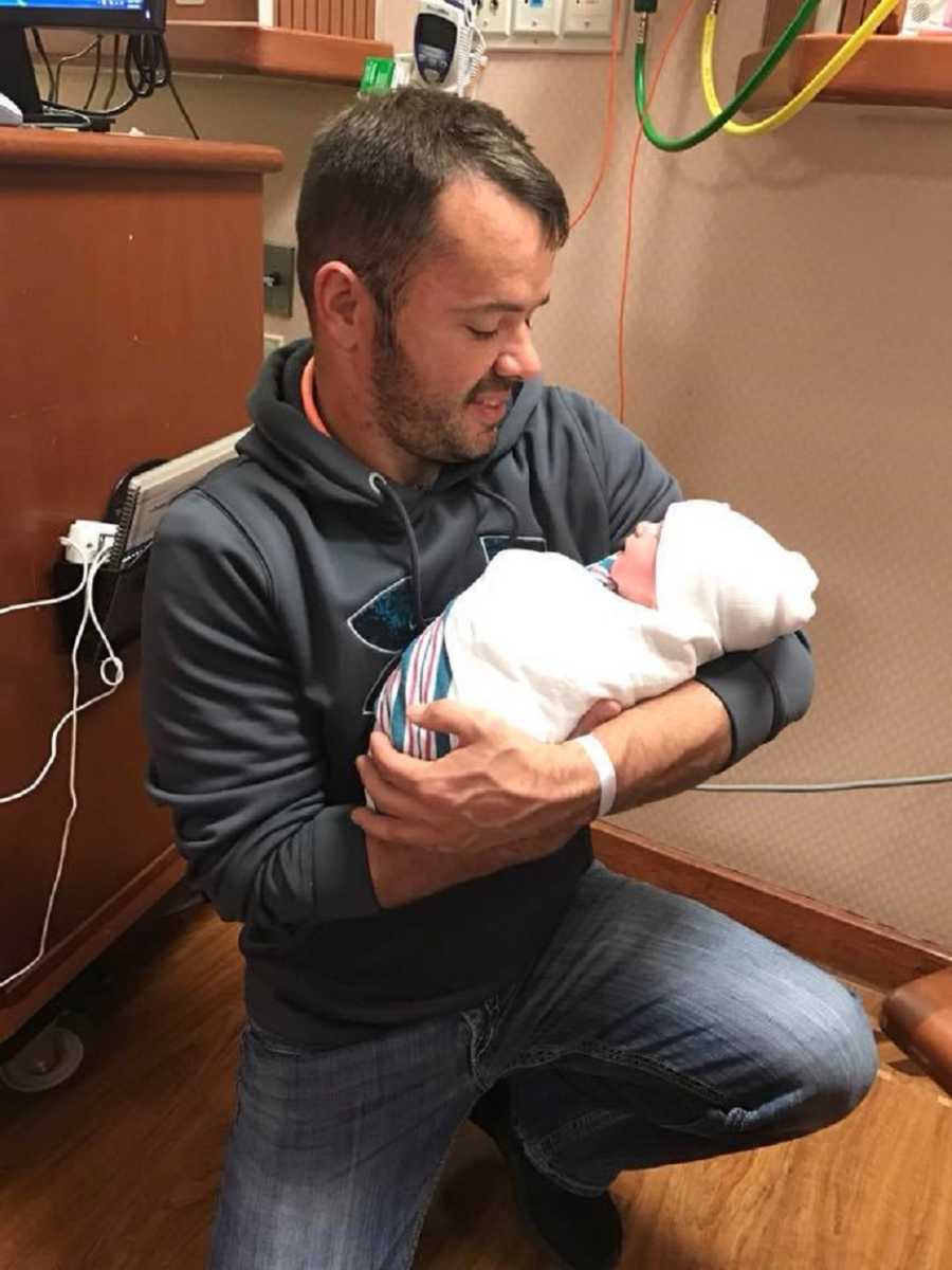 Man who has since passed away from heart attack crouches down on hospital floor holding newborn baby