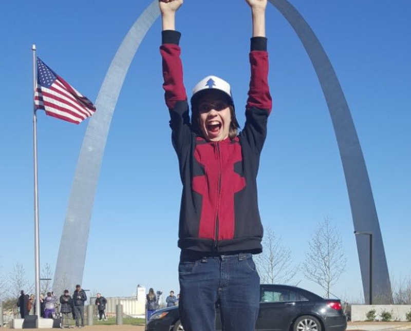 Young boy with ADHD stands smiling arms in the air in font of Gateway Arch
