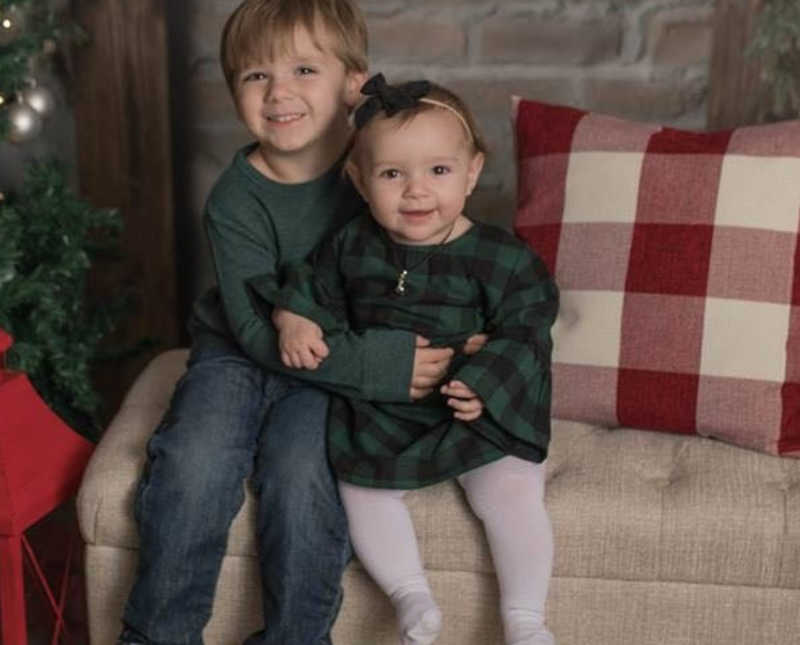 Little boy smiles as he sits on bench in home beside little sister and Christmas tree