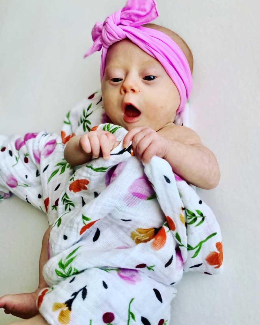 Baby with down syndrome holds floral blanket that is wrapped around her 