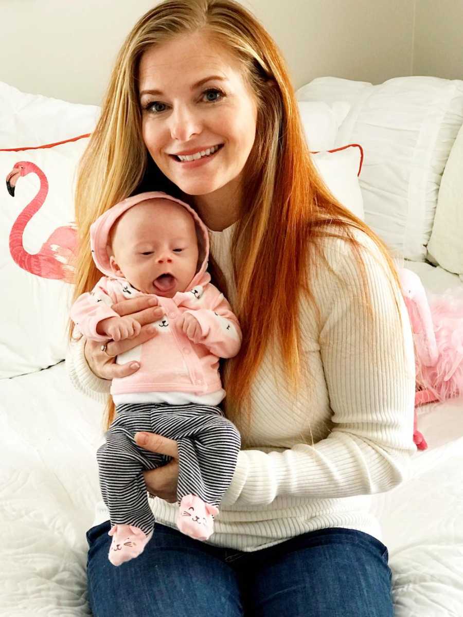 Mother smiles as she sits on edge of bed holding baby with down syndrome