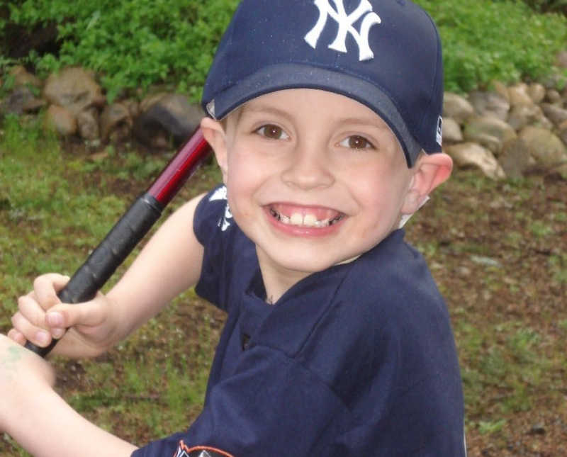 Little boy with ADHD stands smiling in baseball uniform holding bat