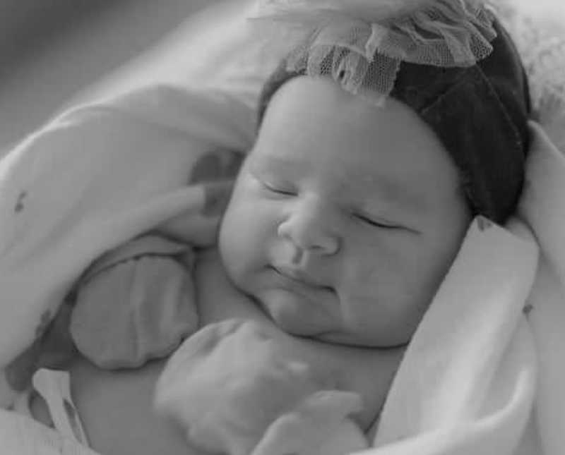 Newborn baby lays asleep with hat on and mittens on her hands