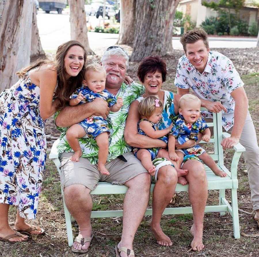 Husband and wife stand on either side of bench where wife's parents sit with her triplet babies in their lap