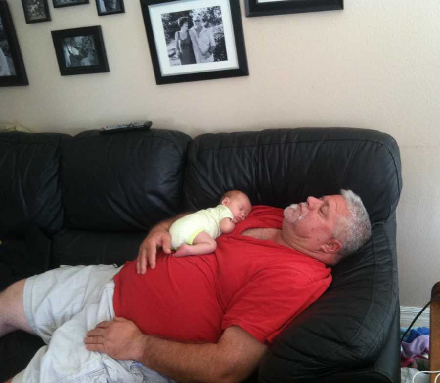 Man lays asleep on couch with baby grandchild asleep on top of him