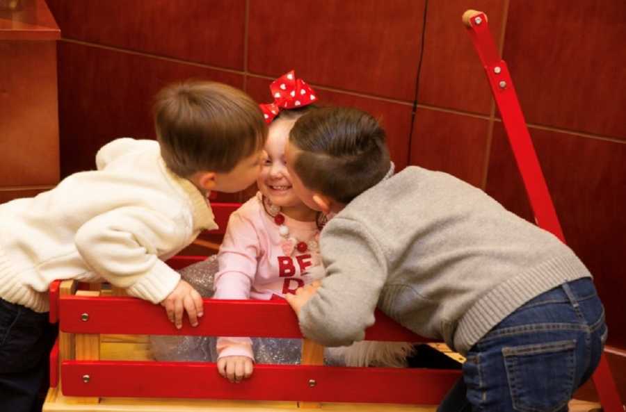 Adopted little girl sits in red wagon in adoption court as two little boys lean over to kiss her