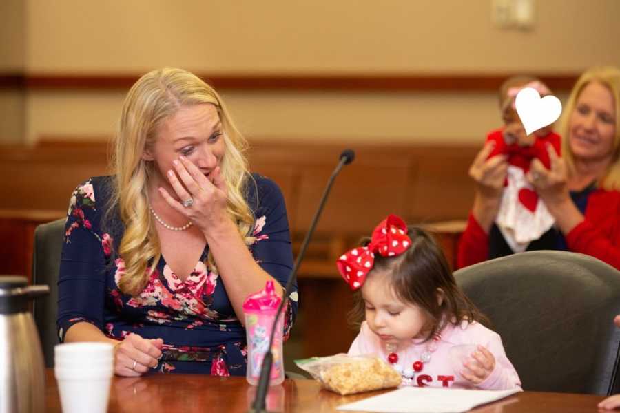 Mother wipes away her tears as she sits at table in adoption court with adopted daughter