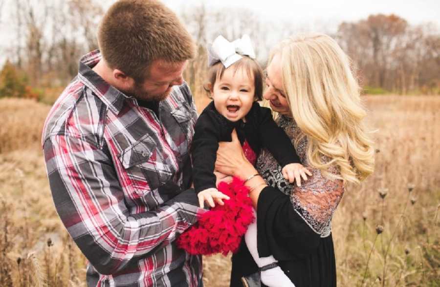 Husband and wife stand in field holding their adopted daughter