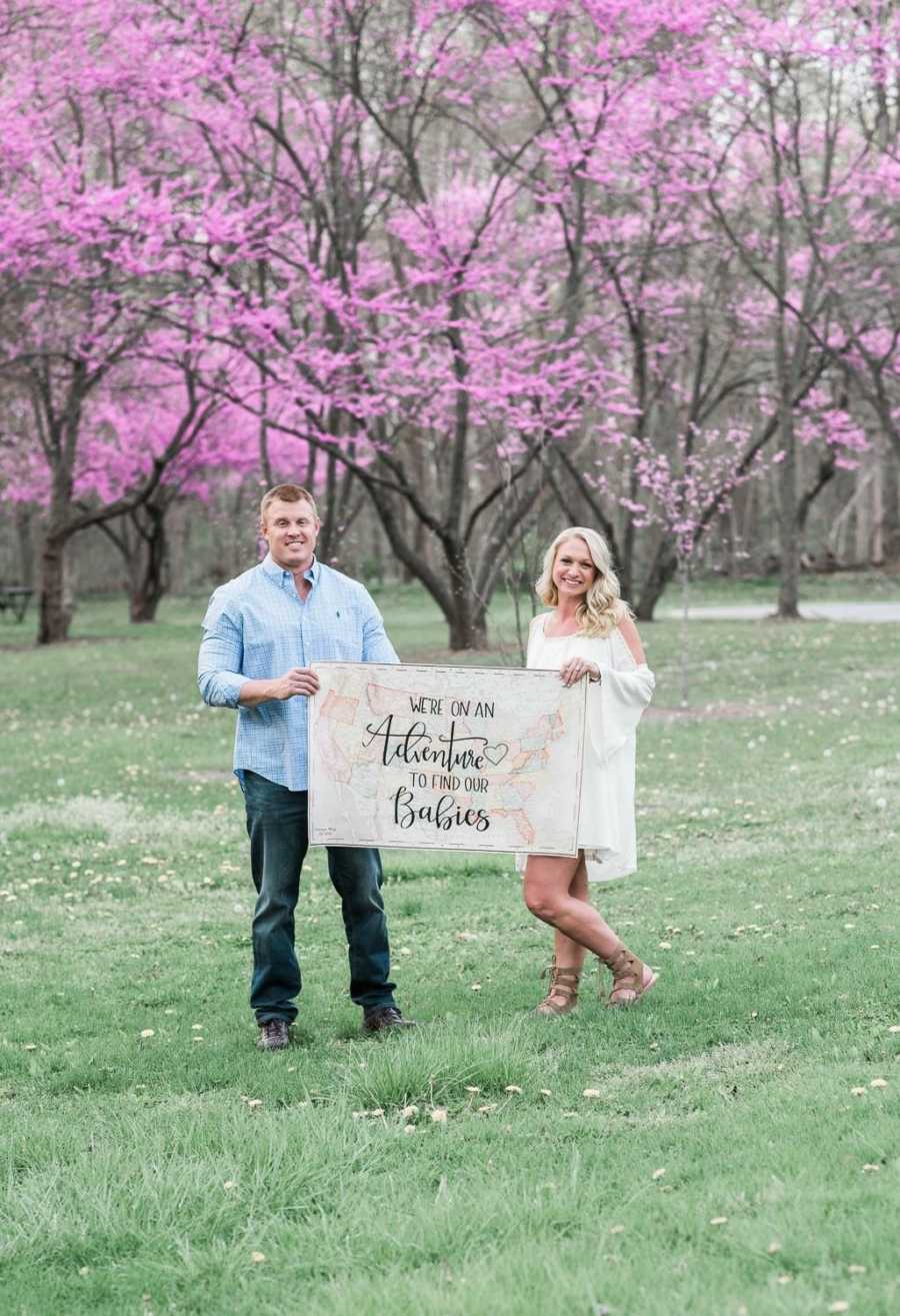 Husband and wife stand outside holding sign that says, "We're on an adventure to find out babies"