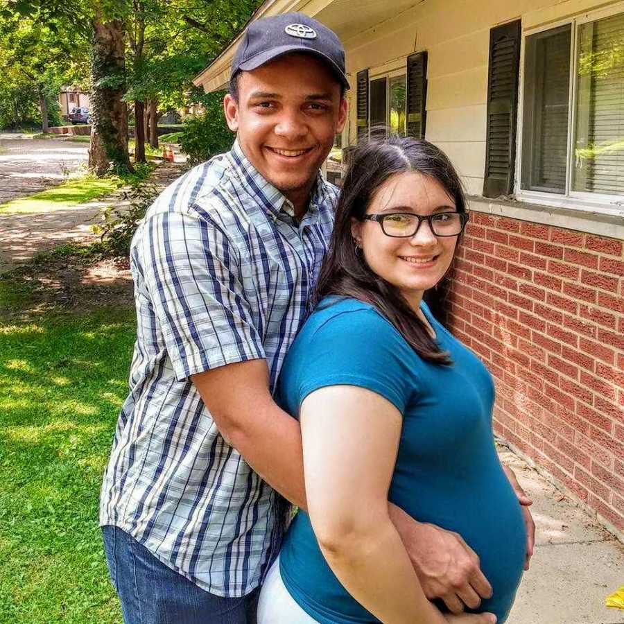 Mixed race husband and wife stand outside of home home as husband holds wife's pregnant stomach
