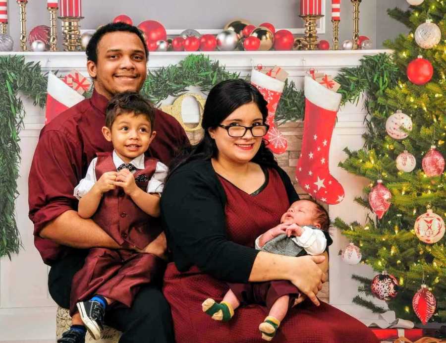 Mixed race husband and wife smile as they sit in front of Christmas backdrop with their two kids