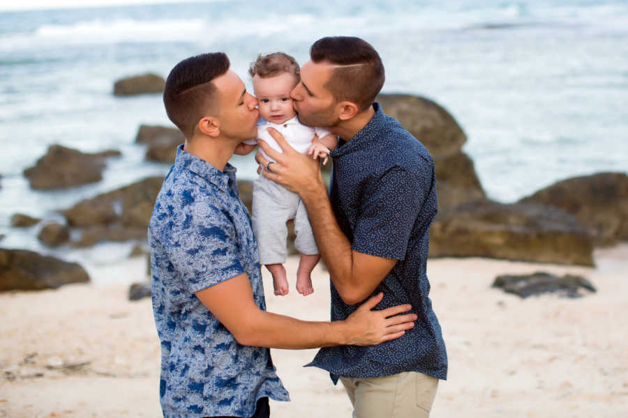 Dads stand on beach holding their son as they kiss his cheeks
