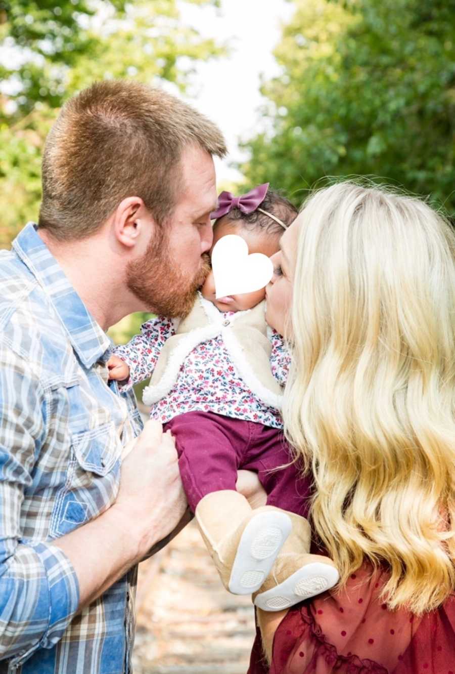 Husband and wife stand outside holding adopted baby as they kiss her cheeks