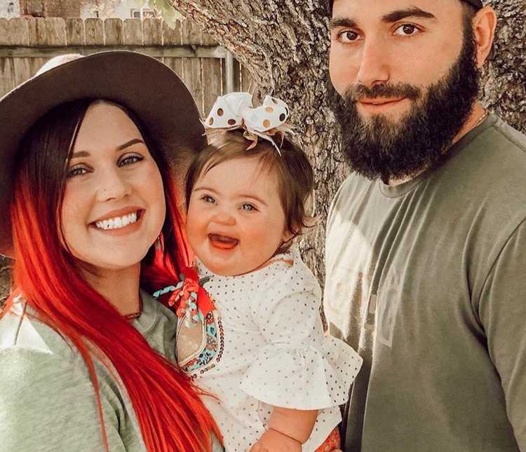 Husband and wife stand smiling outside as wife holds smiling baby with down syndrome