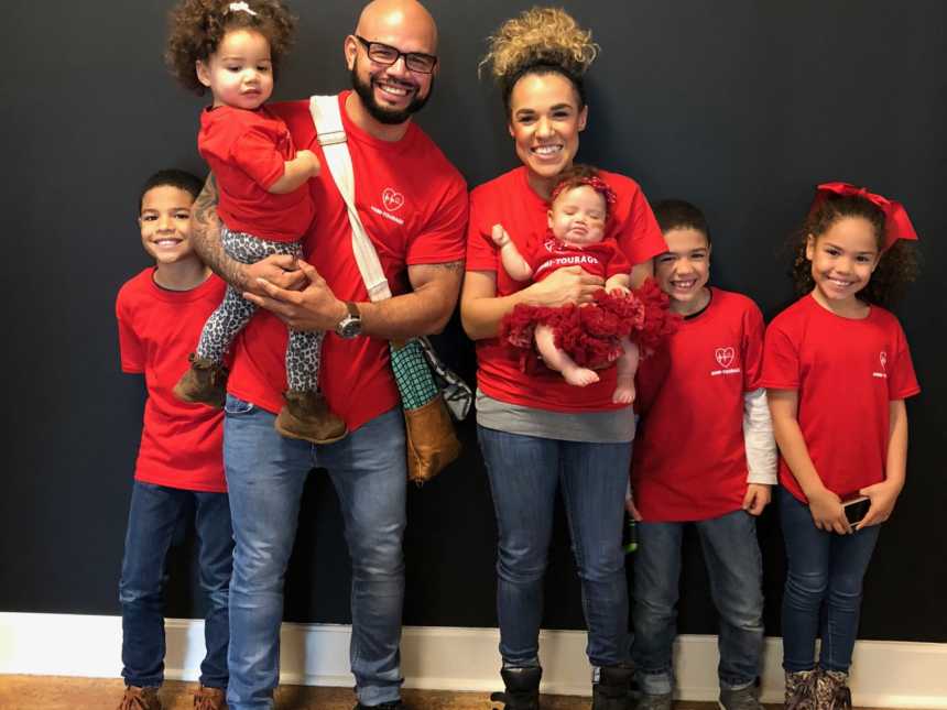 Husband and wife stand in home wearing matching red t-shirts with their five children