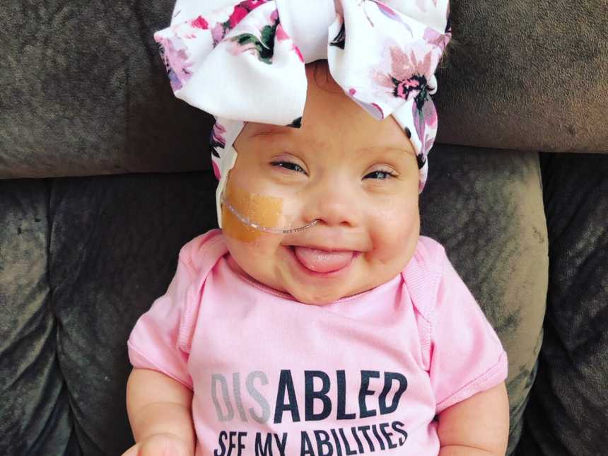 Baby with down syndrome sits smiling in chair with big bow on her head