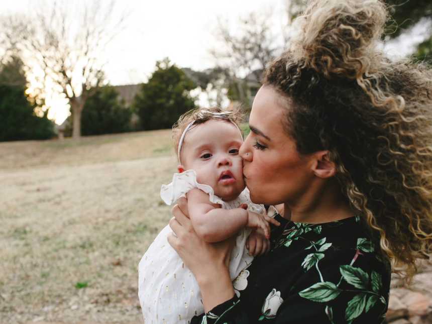 Mother stands outside as she holds daughter with down syndrome and kissing her cheek