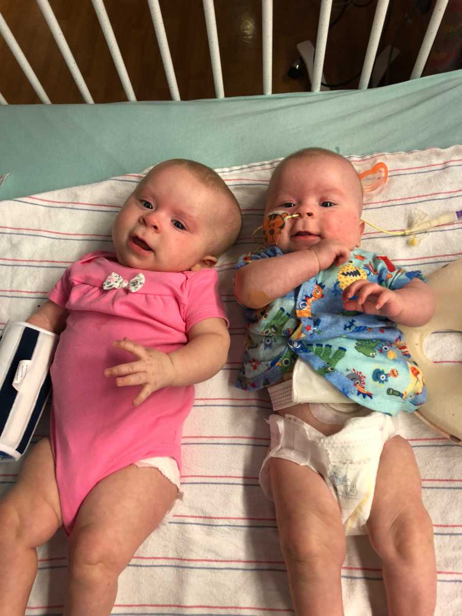 Twin baby girls with cancer lay on backs beside each other in crib in hospital