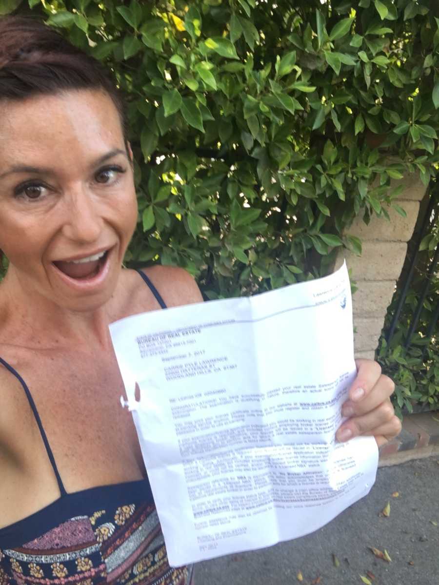 Woman smiles with mouth open as she stands outside holding real estate license in selfie