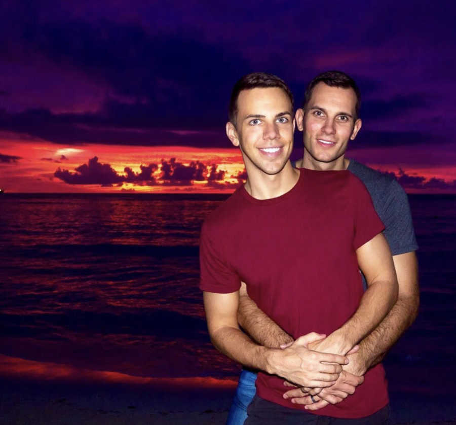 Man stands on beach at sunset with his arms wrapped around his husband