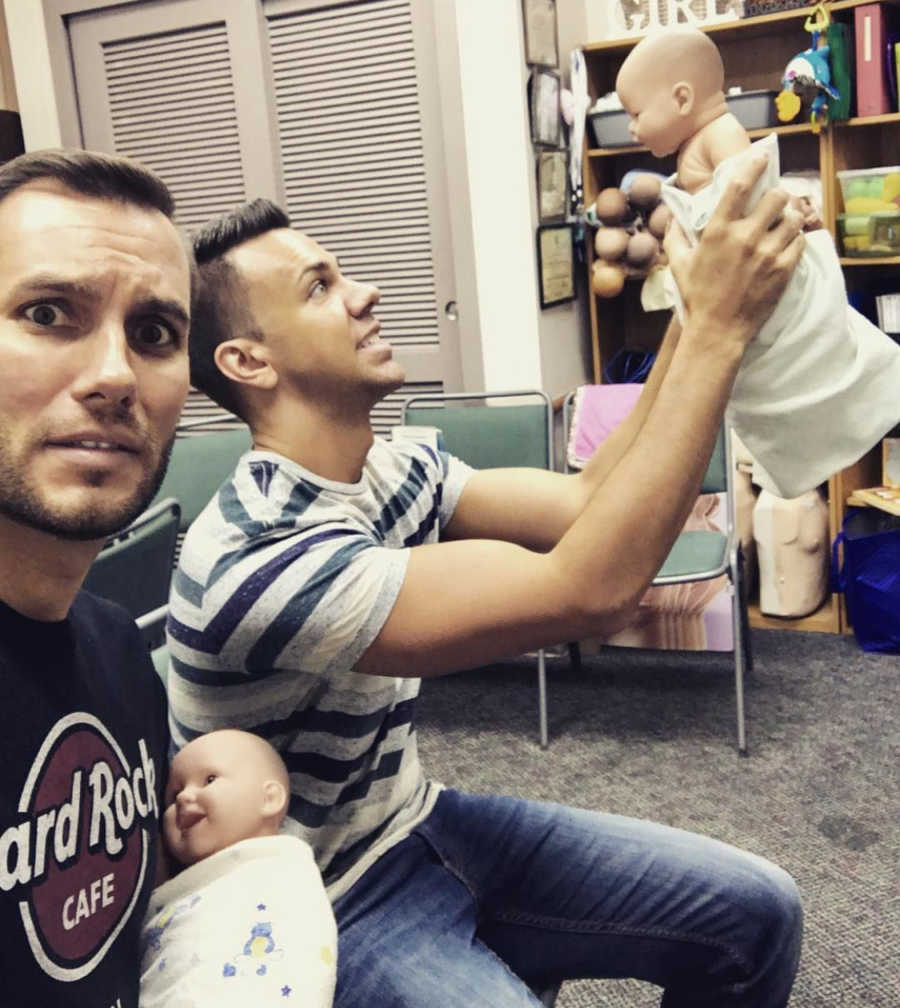 Man takes selfie with scared look on his face as he holds baby doll and husband holds one in the air