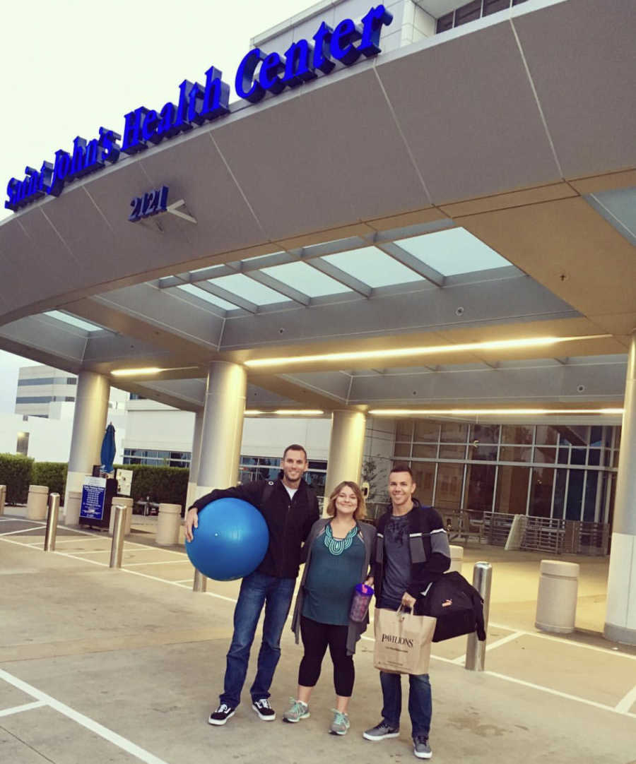 Surrogate stands outside of hospital with father's of the baby on either side of her