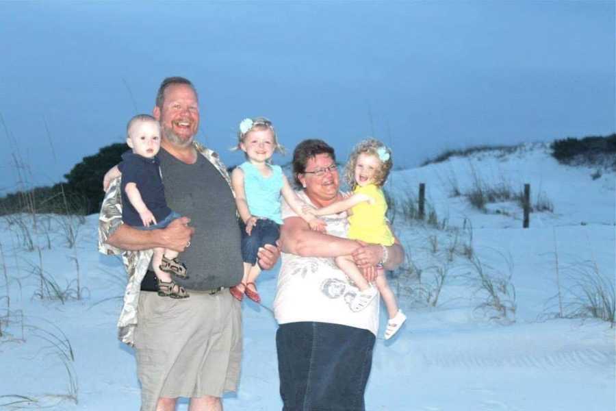 Woman with fibromyalgia stands on beach holding granddaughter as she stands beside her husband who holds to other grand children