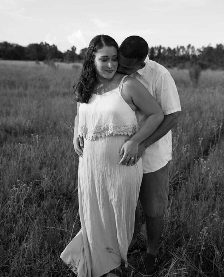 Pregnant woman stands in field as her husband stands behind her with his hands on her hips