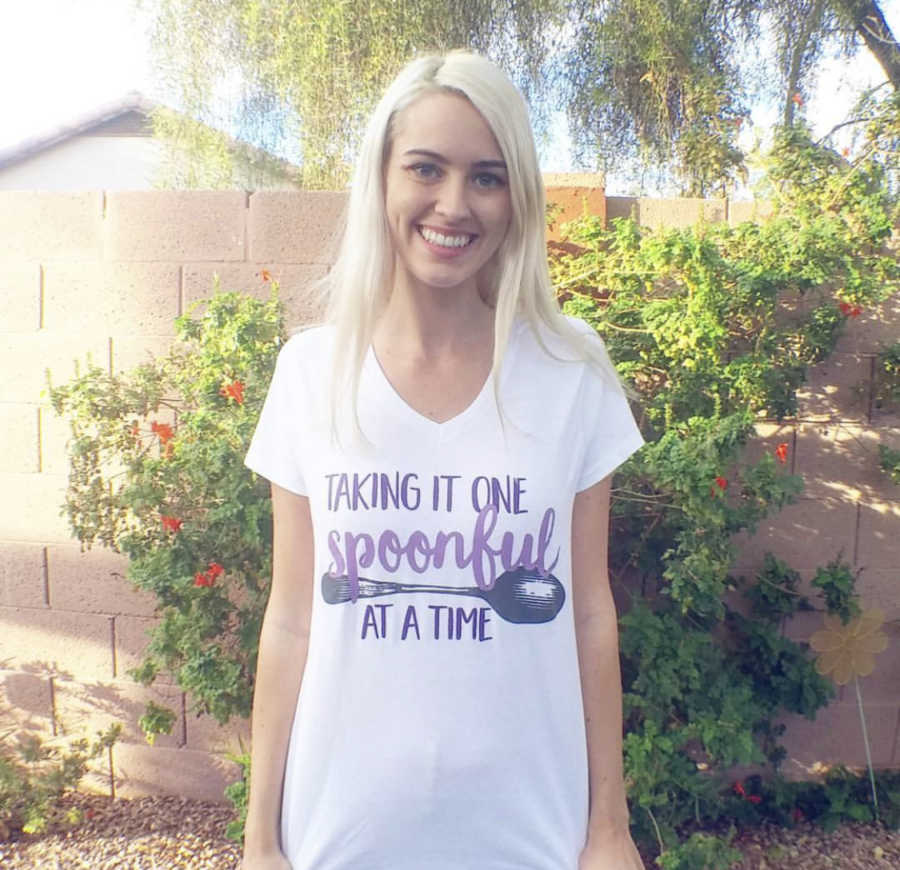 Young woman with POTS stands outside smiling wearing shirt that says, "taking it one spoonful at a time"