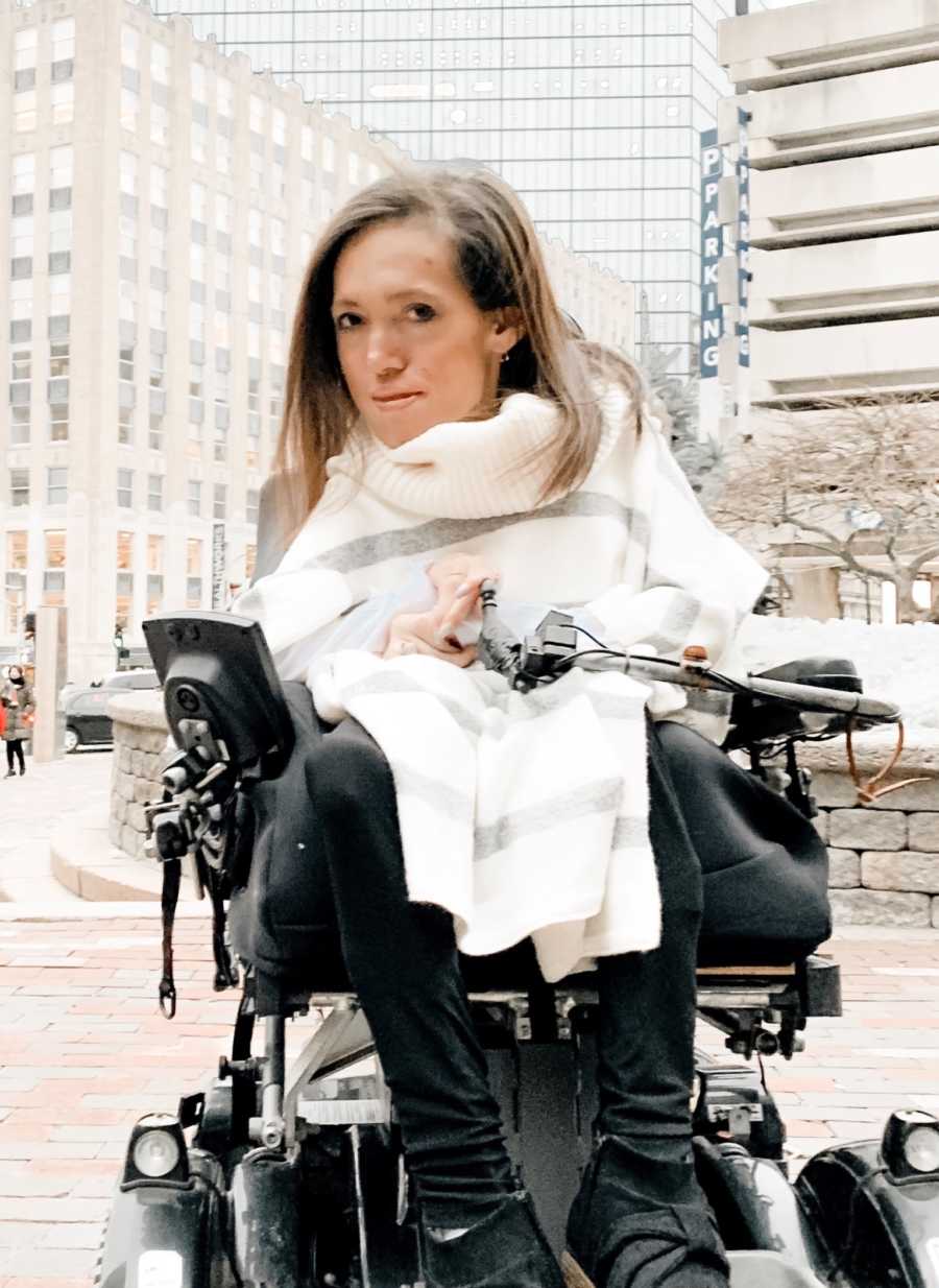 Woman with Spinal Muscular Atrophy sits outside in city in wheelchair 