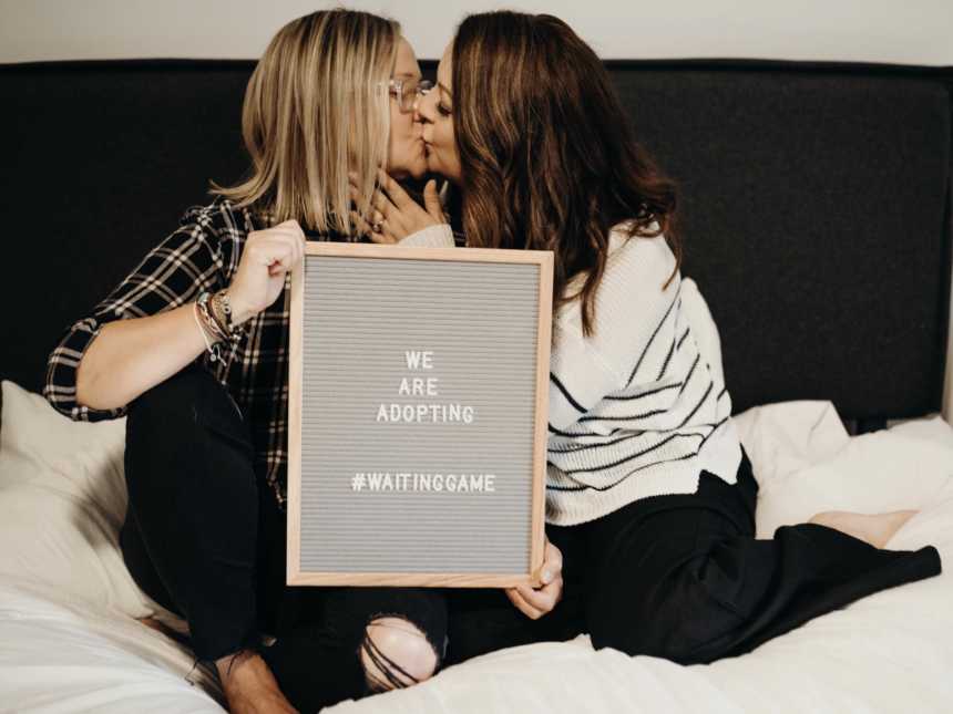 Wife sits in bed beside wife kissing her as she holds sign that says, "We are adopting"