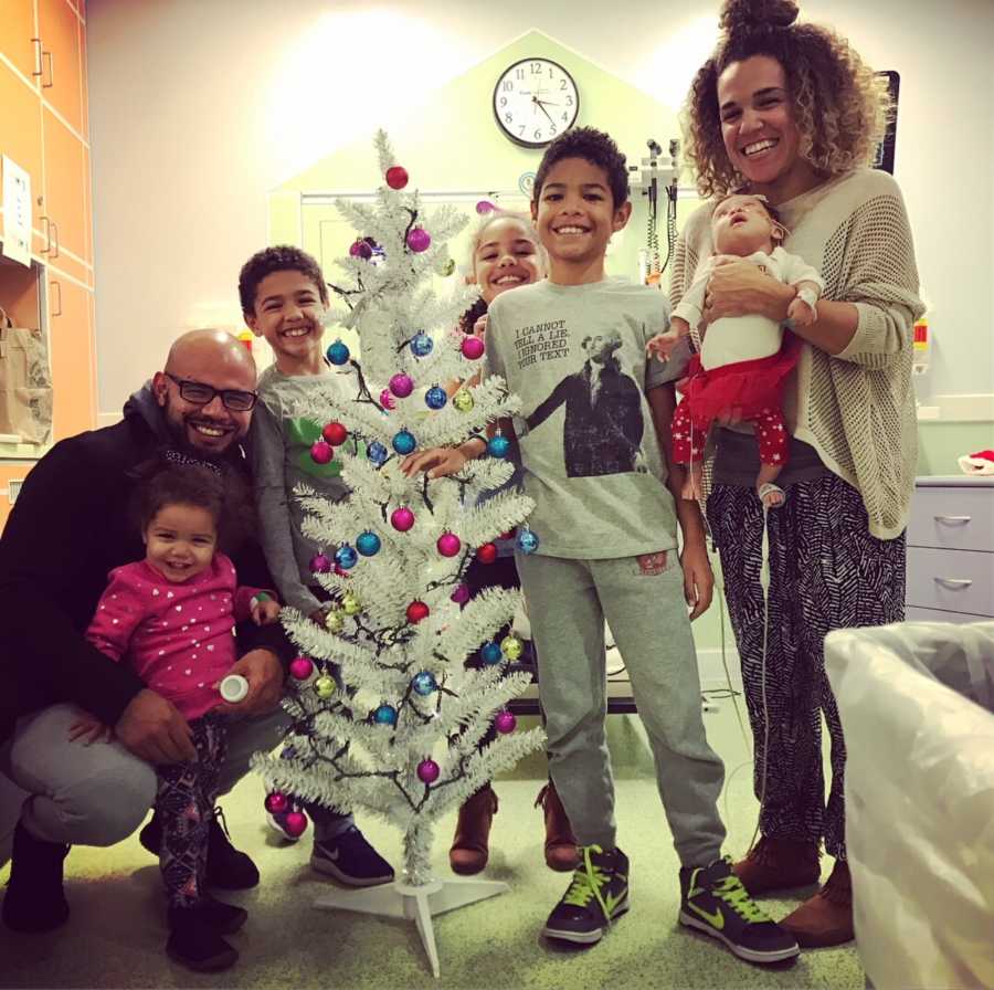 Husband and wife stand in hospital with their five children beside fake white Christmas tree