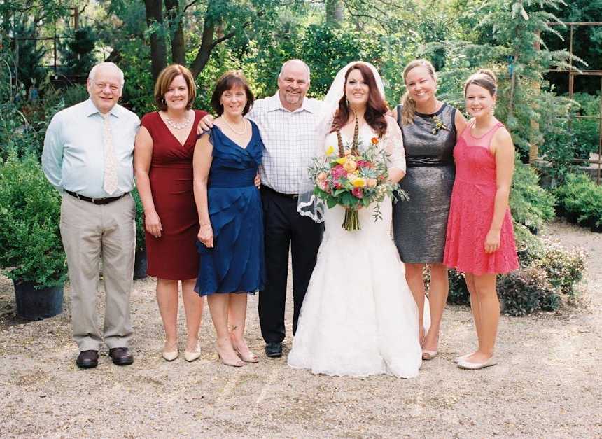 Bride stands smiling outside holding bouquet of flowers beside family members