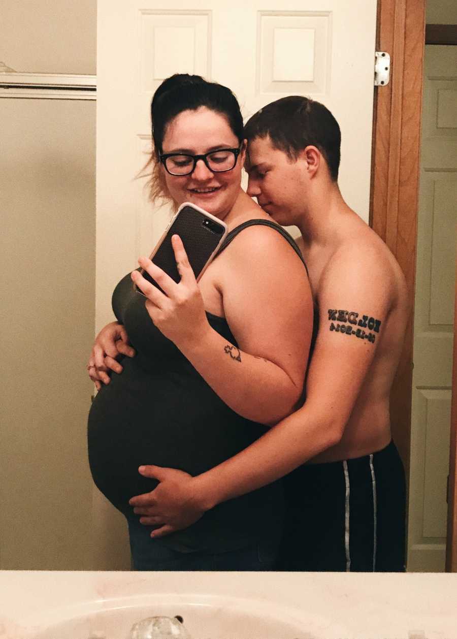 Pregnant woman stands in bathroom taking mirror selfie as husband stands behind her holding her stomach