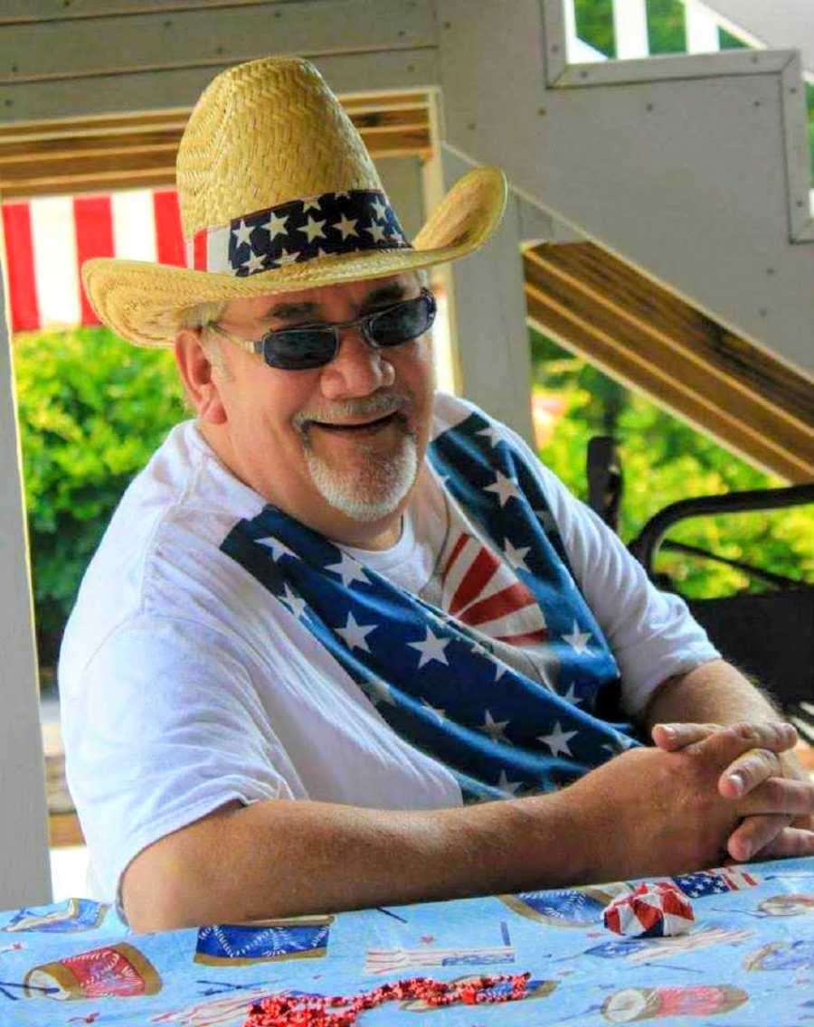 Man smiles as he sits at picnic table wearing American flag t-shirt and straw hat