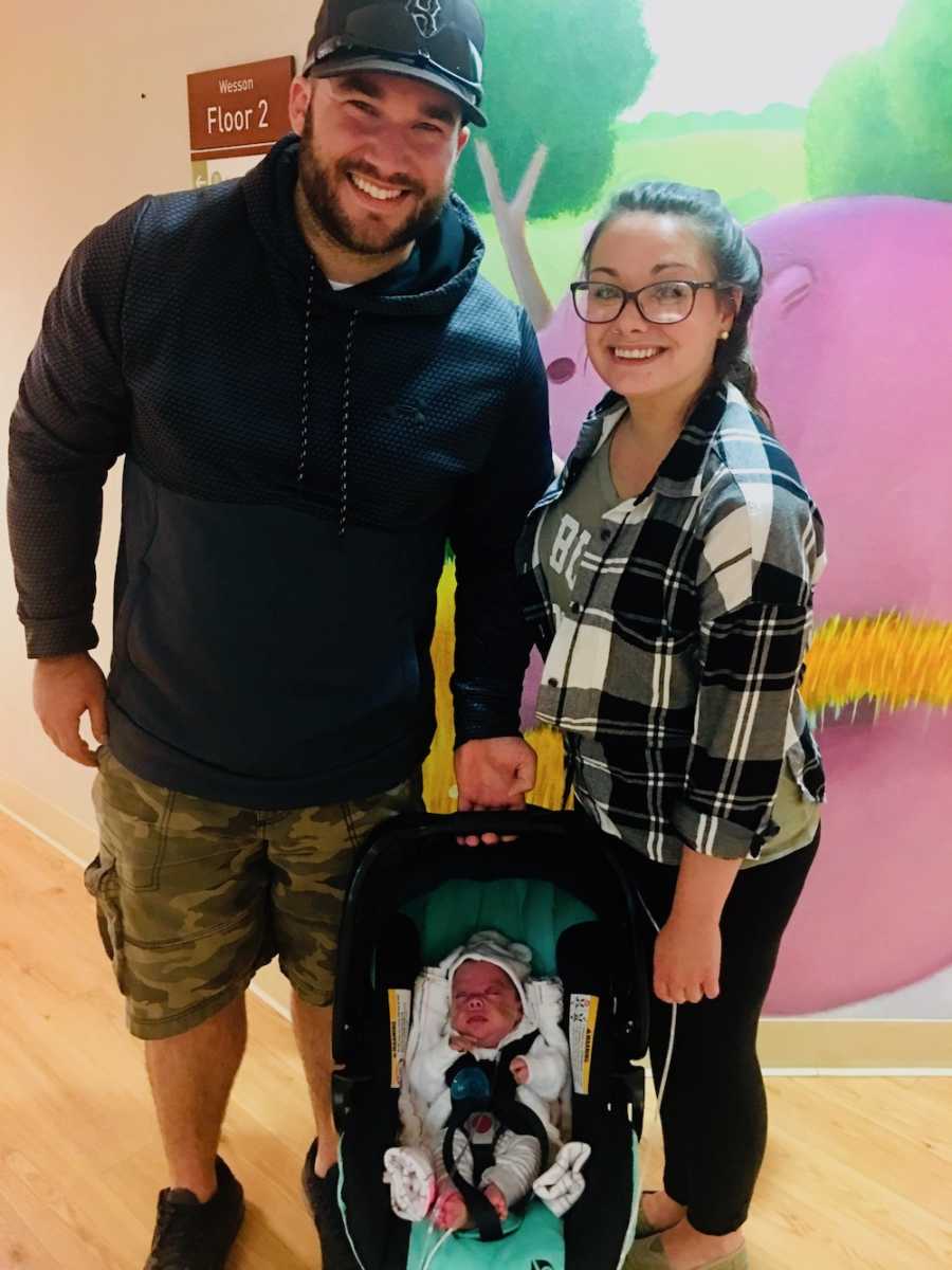 Husband and wife smile as they stand in hospital hallway holding car seat with their baby 