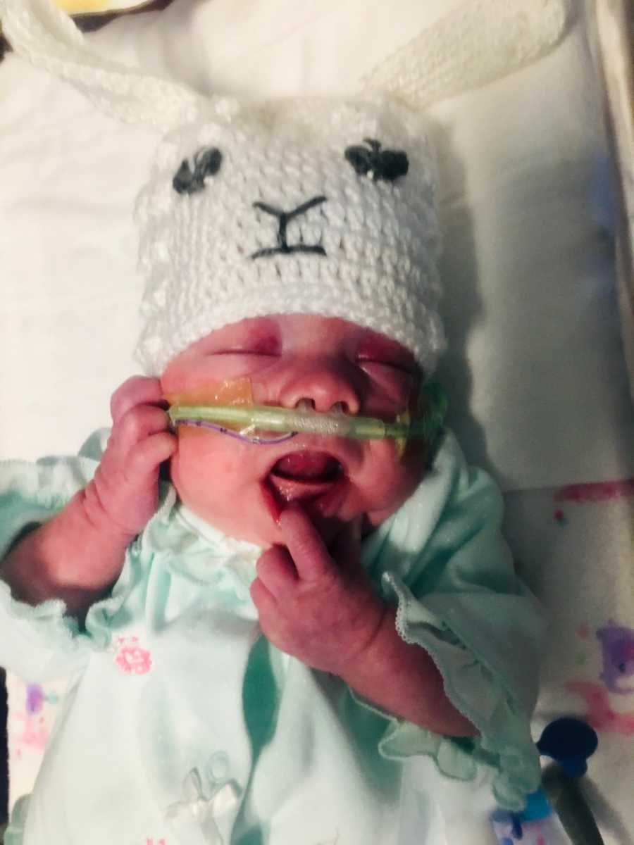 Intubated preemie lays in NICU with finger on her bottom lip and wears white knit hat with face on it