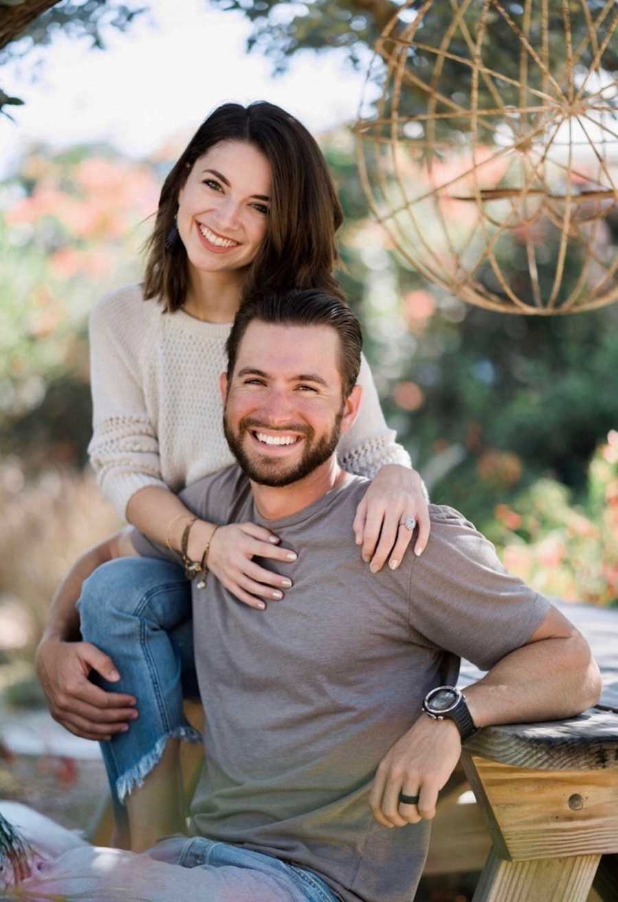 Man smiles on bench of picnic table as wife sits on top of table with hands on husband's shoulder