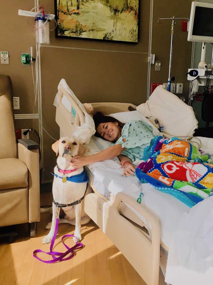 Woman who had spinal tumor lays in hospital bed petting dog that sits beside the bed