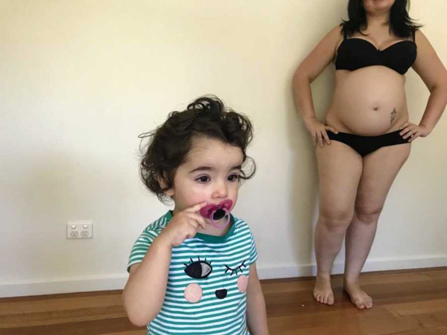 Little girl stands in home with pacifier in her mouth as pregnant mother stands behind her in bra and underwear